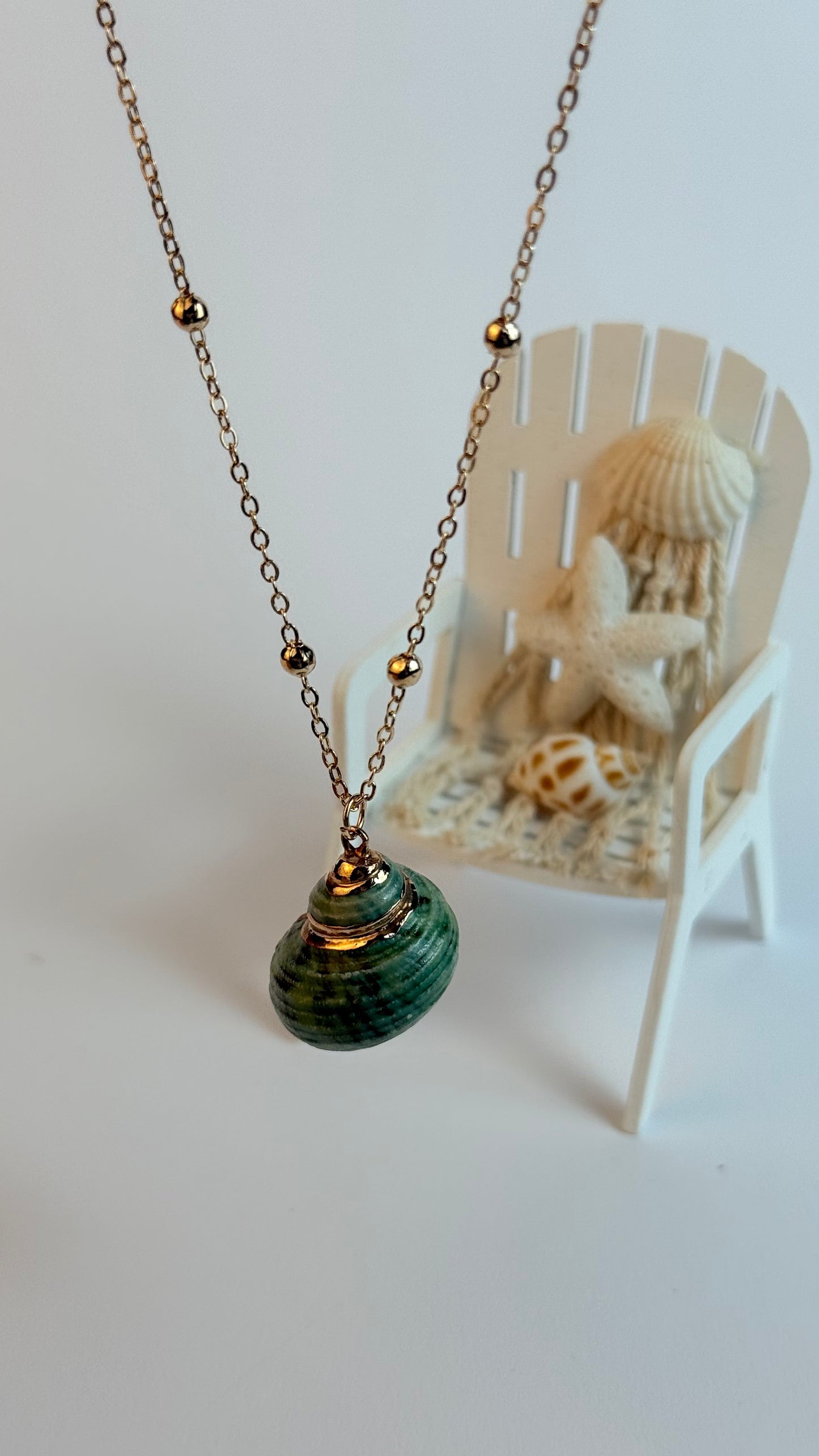 Necklace with a green pendant on a white surface, with a small chair and shells in the background.