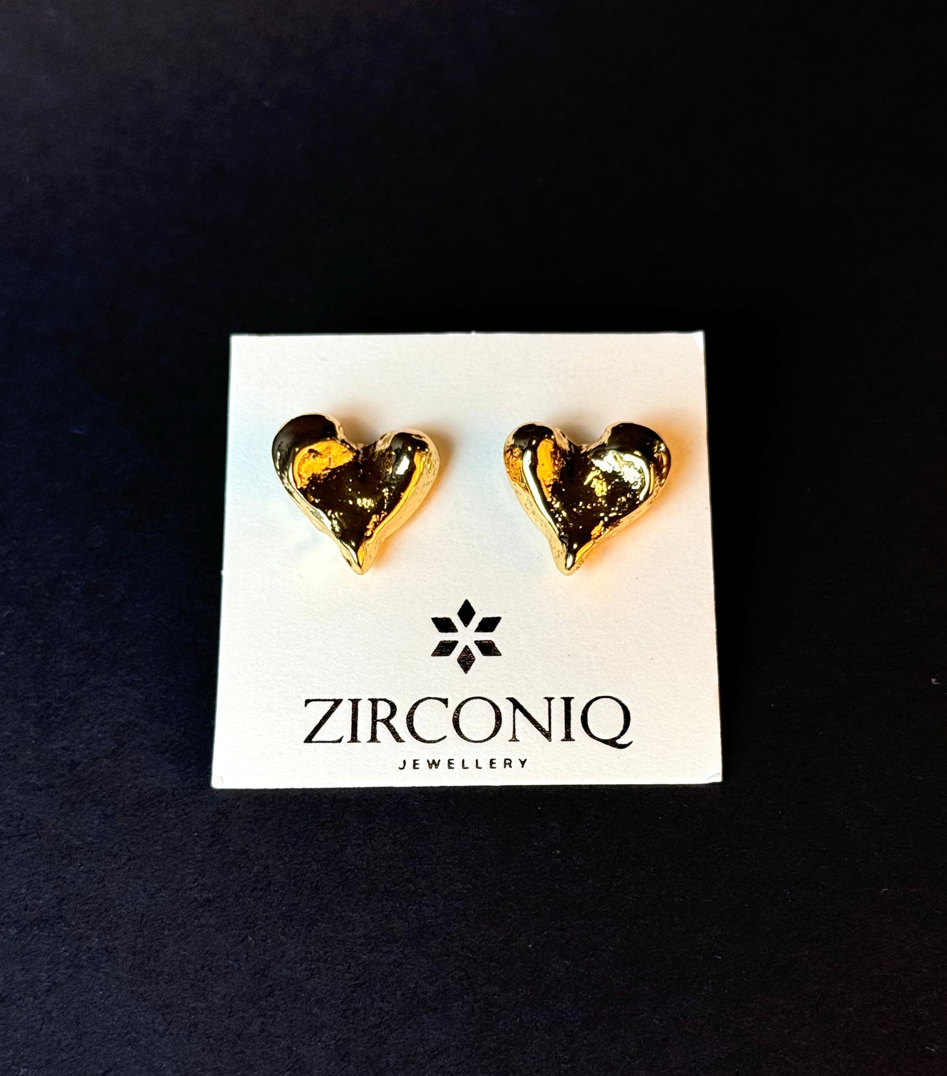 Heart-shaped gold earrings on a Zirconiq branded card against a black background
