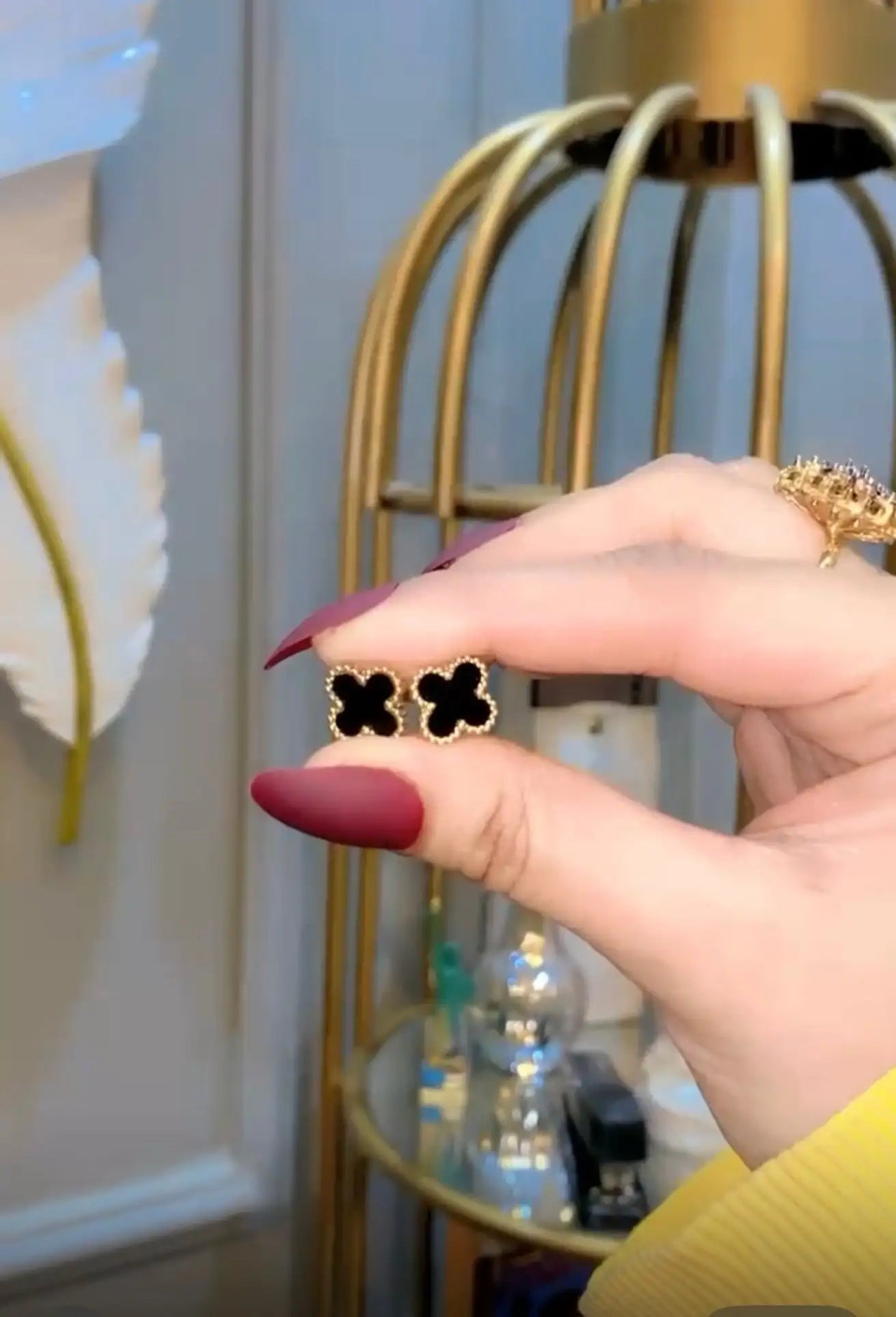 A pair of elegant black four-leaf clover stud earrings with a gold-tone metallic border, delicately held between fingers.